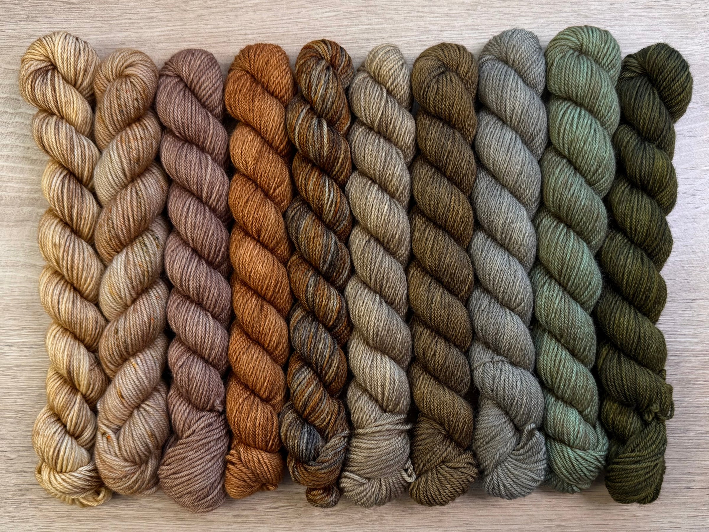 Nine skeins of twisted yarn arranged side by side on a light wood surface, showing a gradient of earthy colors from pale tan and beige to medium brown, rust orange, olive green, and dark forest green.